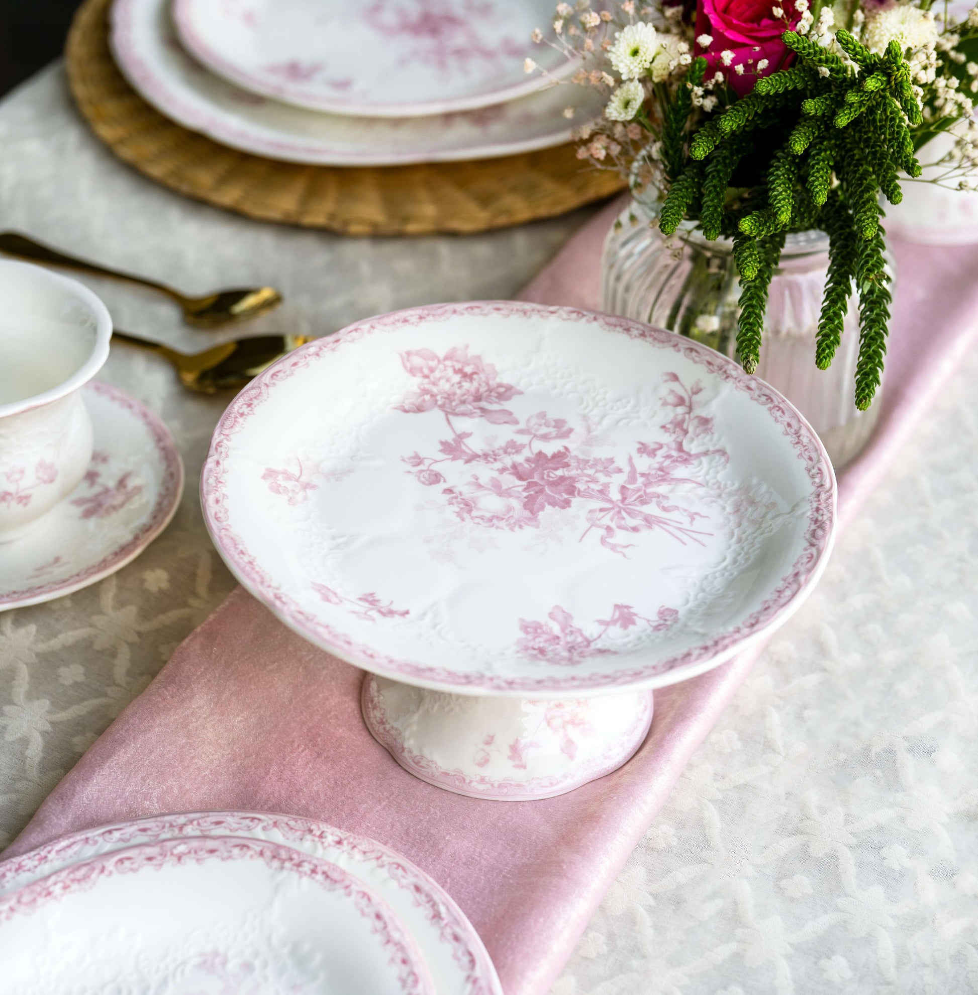 A styled tabletop setting featuring white ceramic plates and a pedestal plate with a delicate pink floral toile pattern, arranged on a pink napkin with gold-toned cutlery and a vase of flowers in the background.