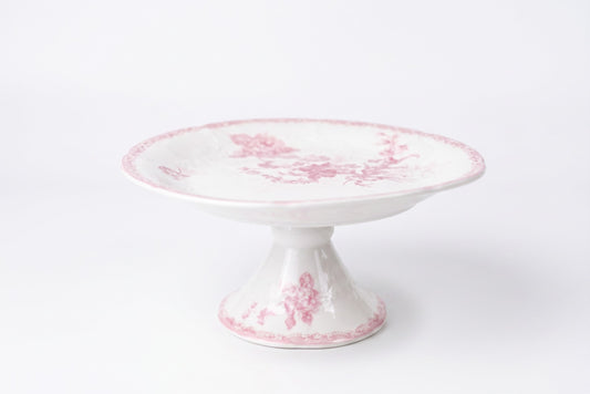 A white ceramic pedestal cake stand with a delicate pink floral toile pattern, set against a plain white background.
