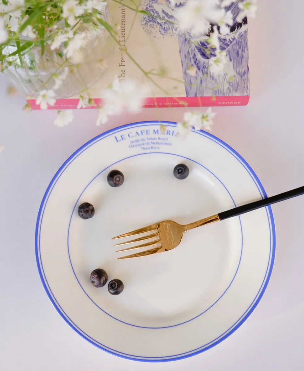 Top-down view of a white bone china quarter plate featuring double blue circular borders and blue "Le Cafe Maria" branding