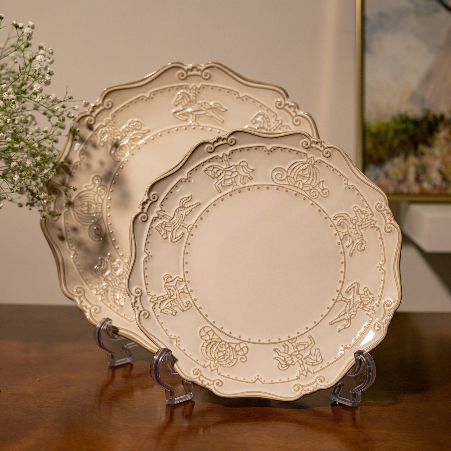 Front-facing view of a 10-inch dinner plate and a 7-inch quarter plate on transparent display stands, both featuring embossed carnival merry-go-round horses and carriage carvings
