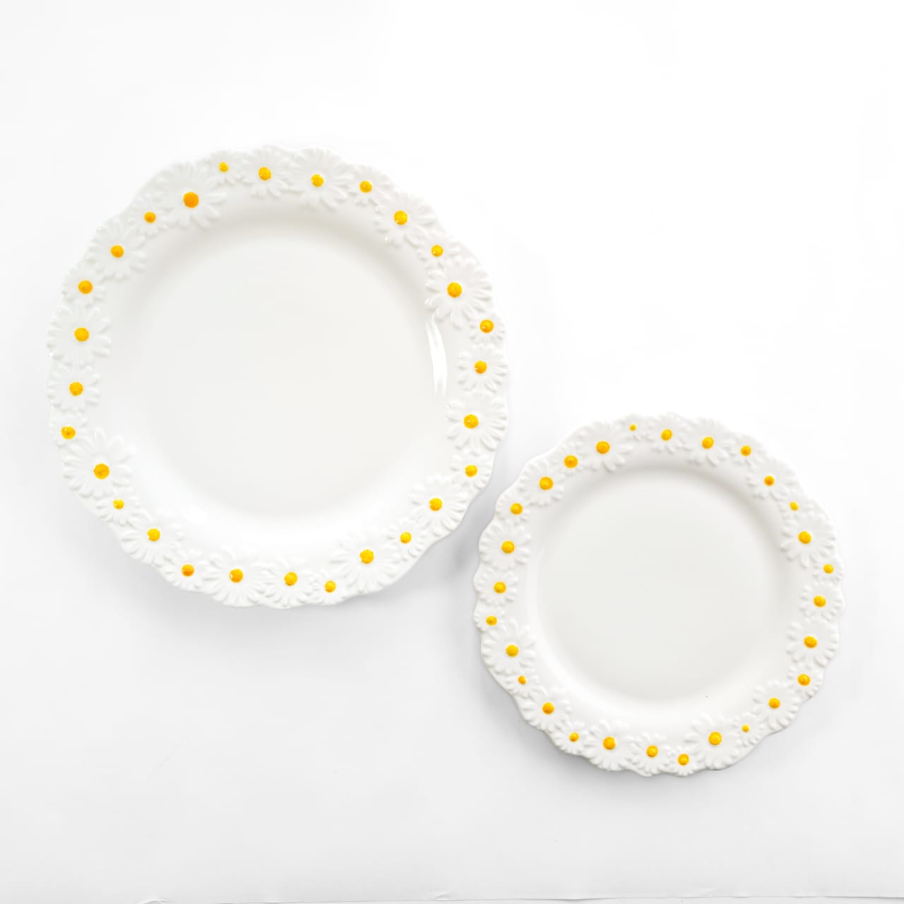 Top-down view of a white bone china dinner plate and a smaller quarter plate, both featuring scalloped edges and embossed daisy patterns with yellow centers