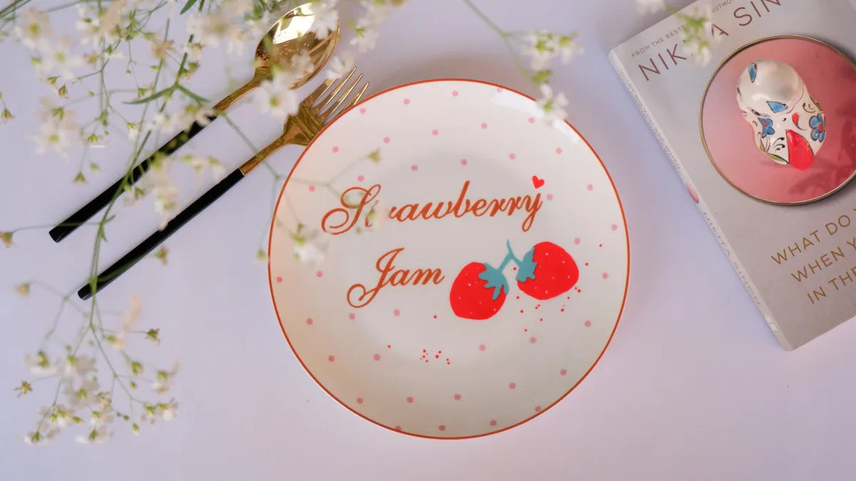 Top-down lifestyle view of an 8-inch white bone china plate featuring red "Strawberry Jam" cursive text and two red strawberry illustrations - Birdy Home Design