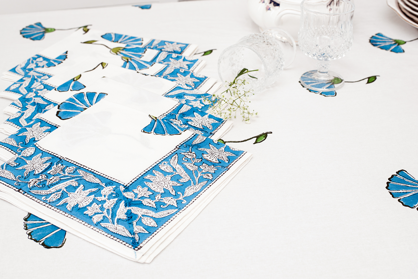 A stack of white cotton napkins with blue floral block print borders placed on a matching white table cloth with blue floral motifs