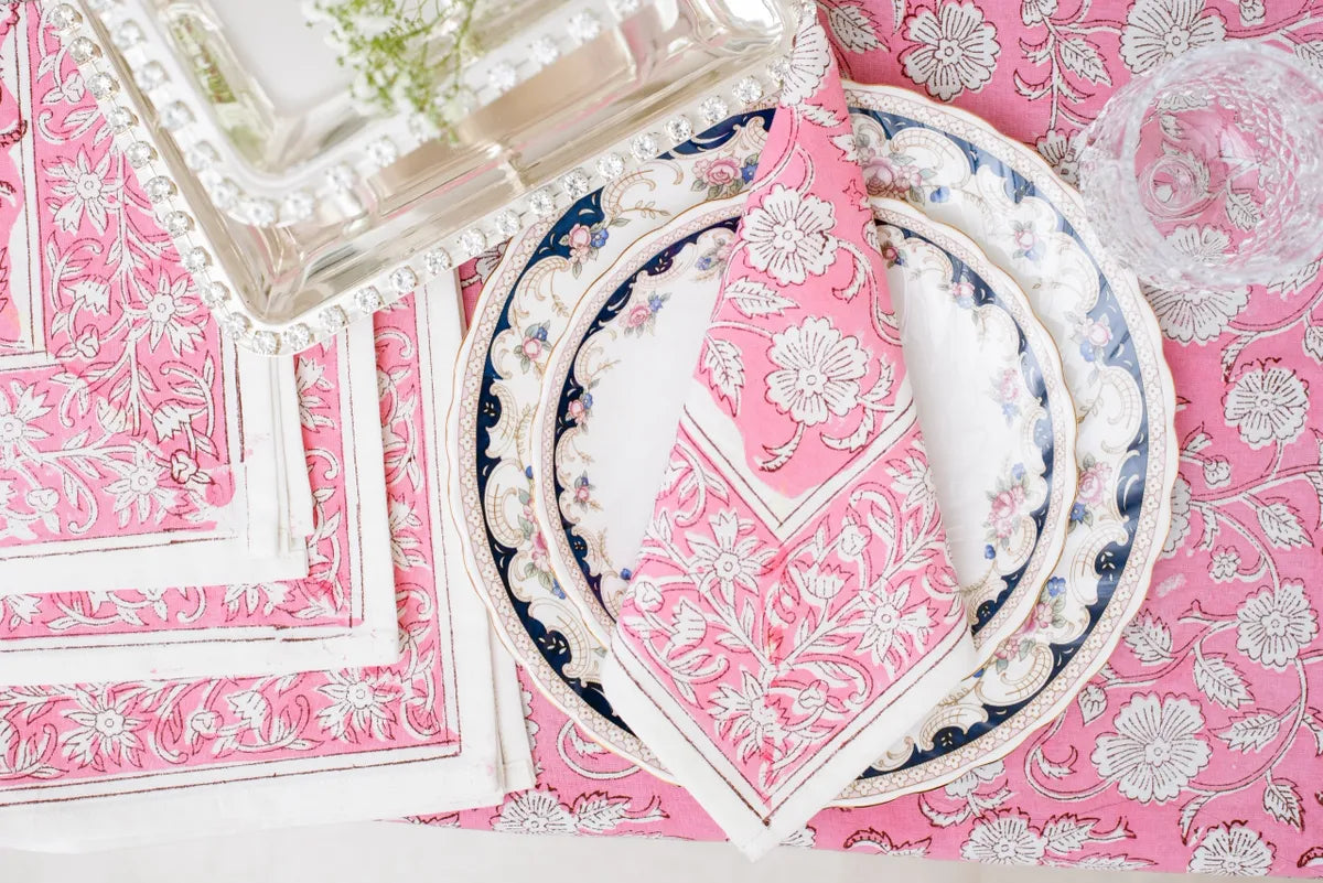 A square pink cotton napkin featuring a white Rajasthani floral block print pattern and a thin white border