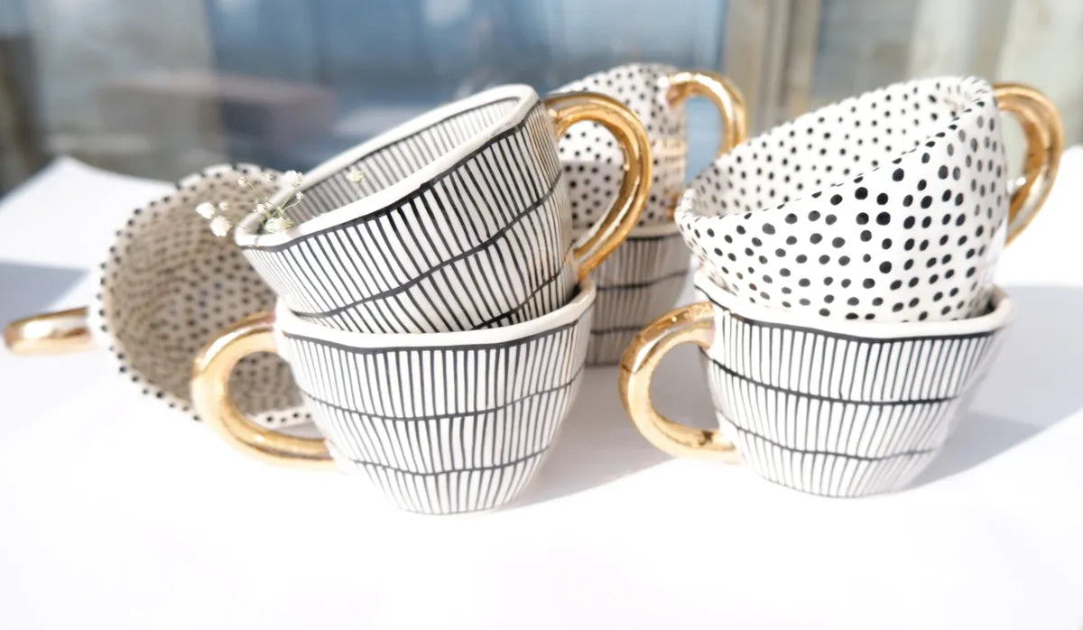 A group of four stoneware mugs featuring various black and white vertical striped and dotted patterns, each with a contrasting metallic gold handle