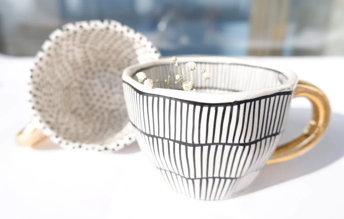 Side-by-side view of two stoneware mugs featuring vertical and dotted black and white patterns with contrasting metallic gold handles