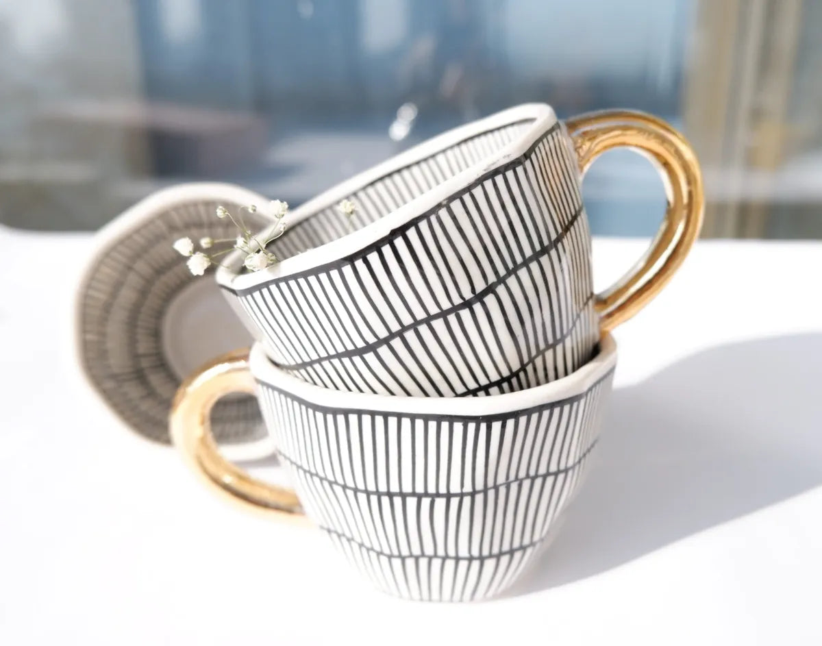 A stacked view of two stoneware mugs featuring black and white vertical striped patterns with contrasting metallic gold handles