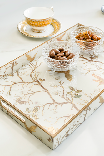 Angled view of a rectangular white wooden tray featuring a gold border and an intricate gold chinoiserie pattern of birds and branches